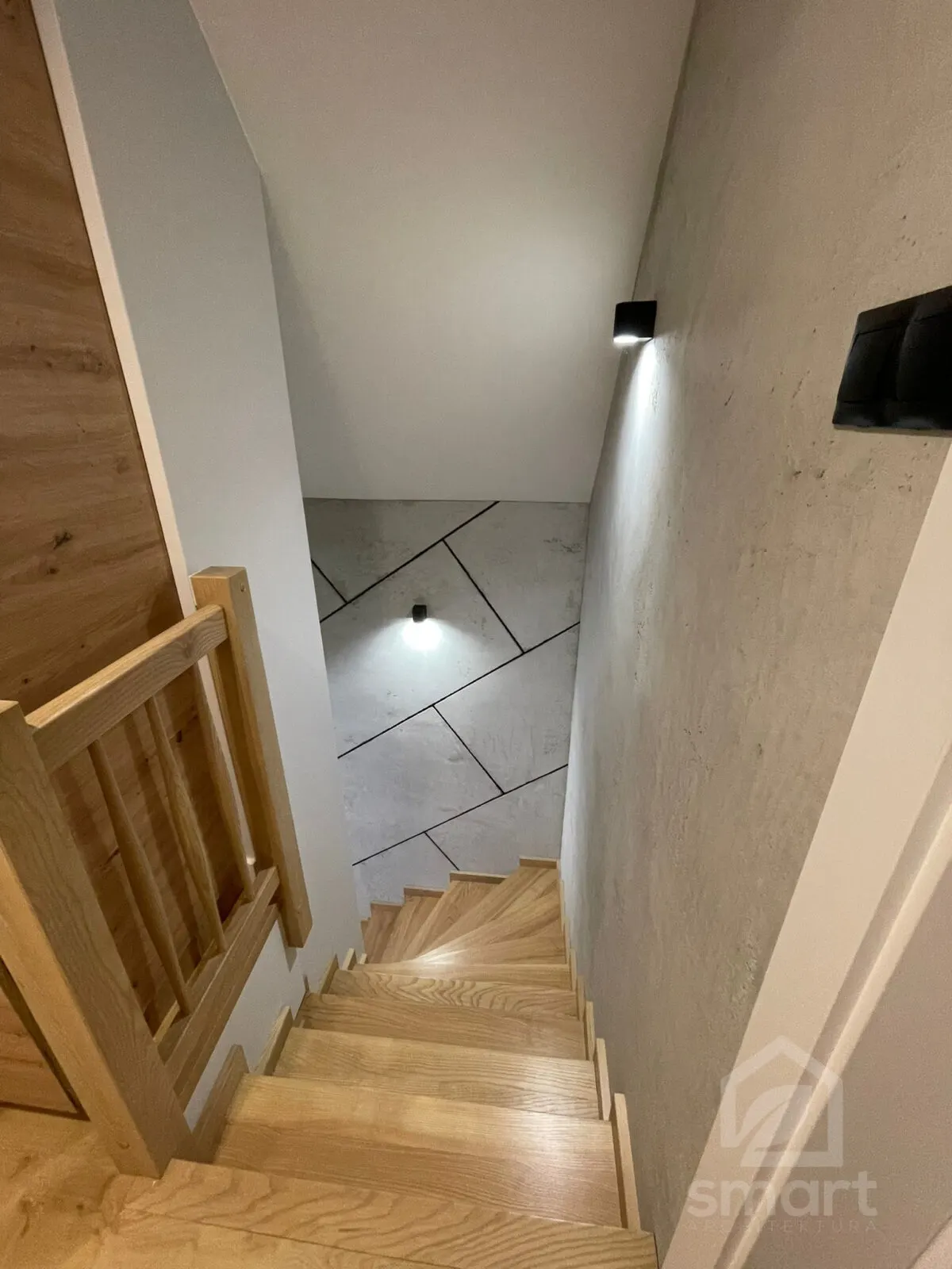 Widok ze schodów na betonową ścianę z geometrycznym podziałem i drewnianą balustradę
