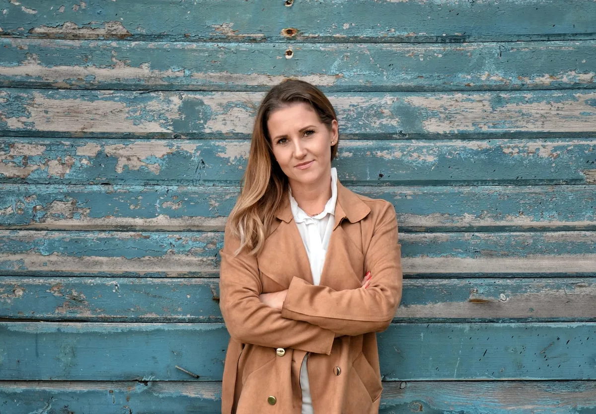 Paulina Łukaszewska w beżowym płaszczu, z założonymi rękami, na tle niebieskiej drewnianej ściany